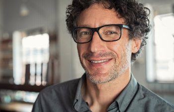 Man with curly hair and glasses smiling indoors, wearing a button-up shirt.