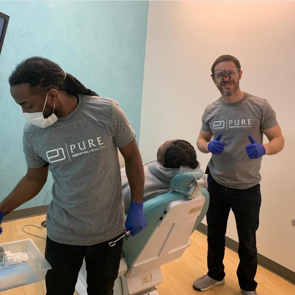 Two dental professionals, wearing gray "Pure Dental Health" t-shirts and blue gloves, stand next to a reclining dental chair with a patient in a clinical setting.