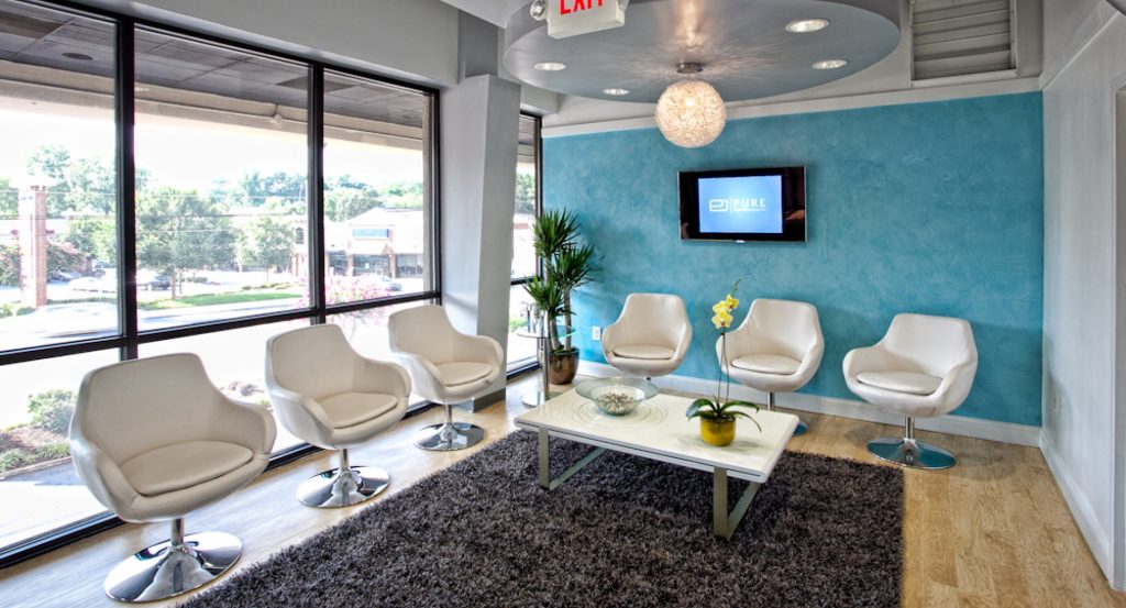 Modern waiting room with white chairs, a central table, a gray rug, and a wall-mounted TV. Large windows provide natural light, and a plant adds a touch of greenery.