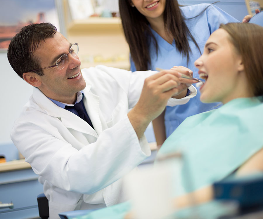 A dentist examines a patient's teeth while seated in a dental chair, assisted by a dental hygienist.