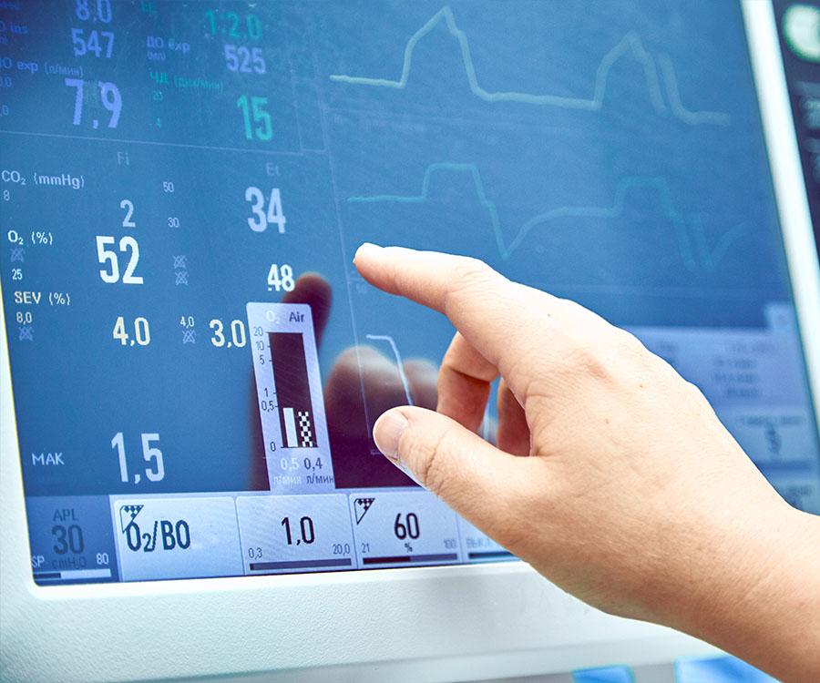 A hand adjusts settings on a medical monitor displaying various vital signs and readings, including heart rate and blood pressure.