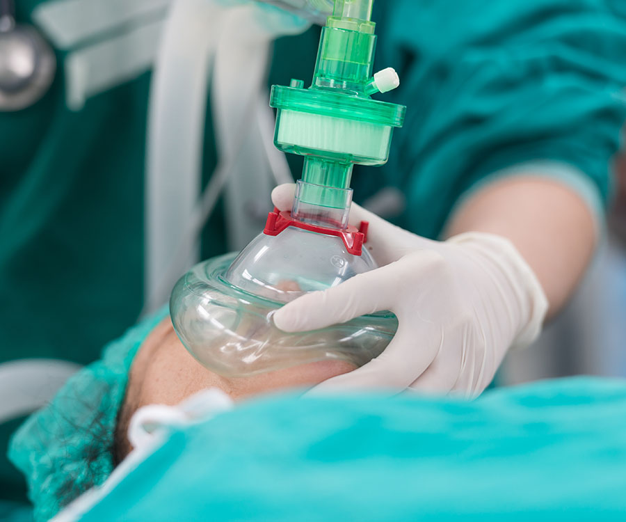 A medical professional in scrubs holds an oxygen mask over a patient's face in a clinical setting.