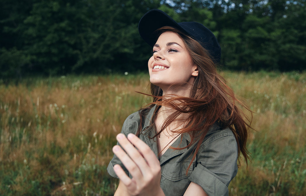 A young woman with long brown hair wears a black cap and green shirt, smiling outdoors in a grassy field with trees in the background.