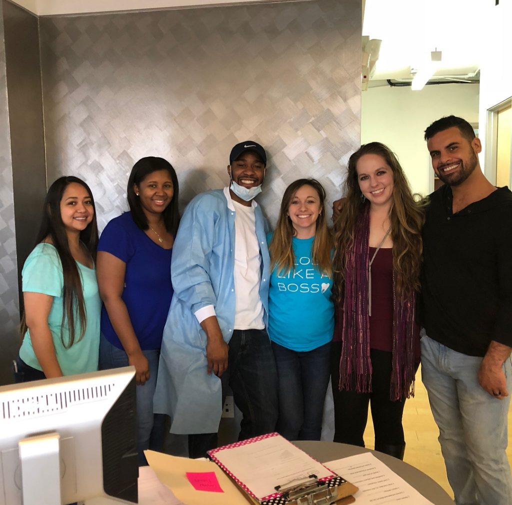 Six people standing together in an office setting, smiling at the camera. One person is wearing a dental uniform. A desk with papers is in the foreground.