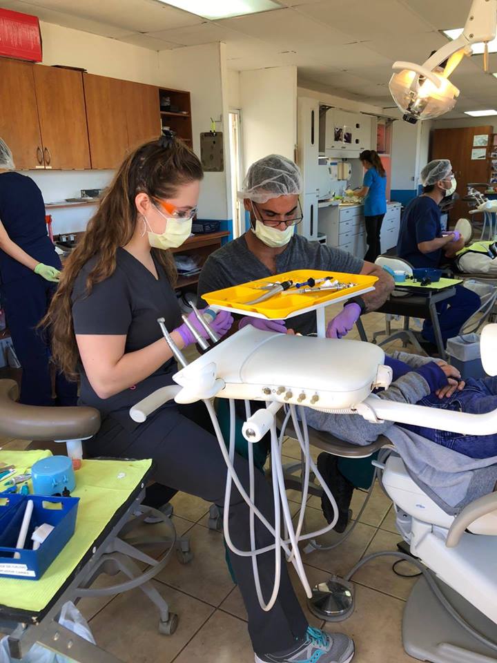 Dental professionals in protective gear work on a seated patient in a clinic setting, with dental tools on a tray.