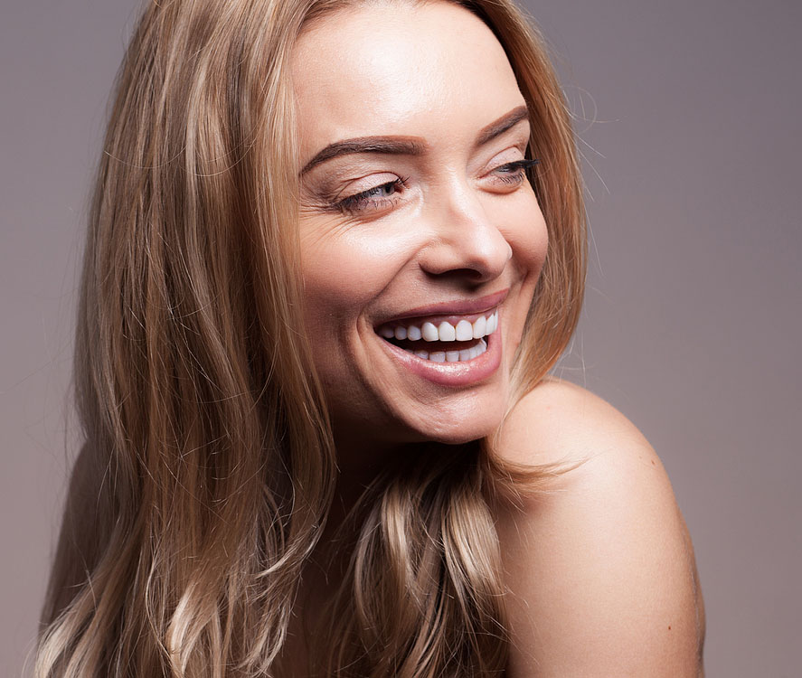 A woman with long blonde hair smiles and looks to the side against a plain background.