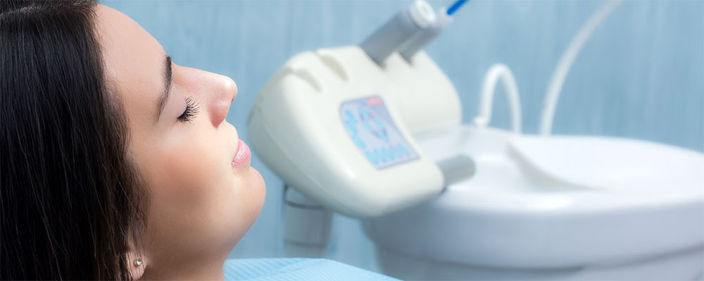 A woman reclines in a dental chair with her eyes closed. Dental equipment is visible in the background.
