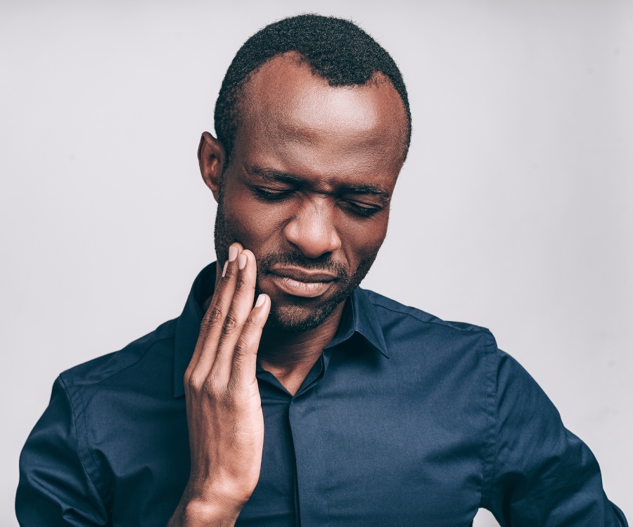 Man in a dark blue shirt touches his cheek and closes his eyes, appearing to experience pain or discomfort.