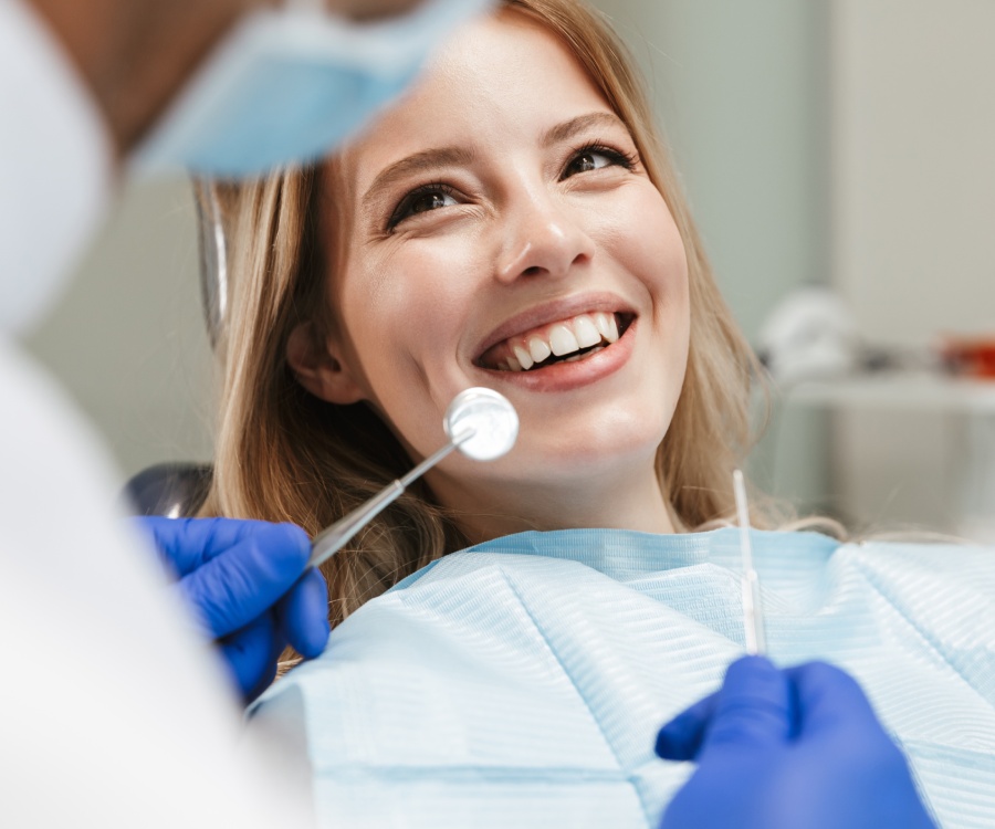 A woman smiles in a dental chair as a dentist wearing gloves holds dental instruments near her mouth.