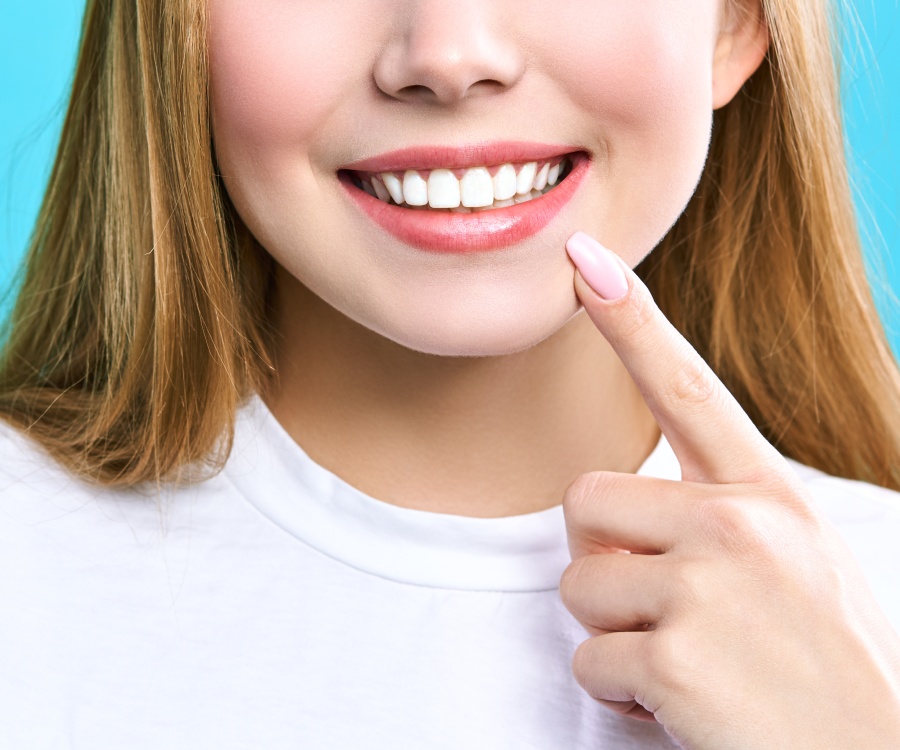 A person with long hair and a white shirt smiles, showing white teeth, while pointing to their mouth against a blue background.