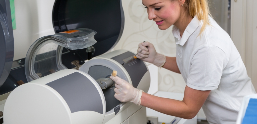 A woman in a lab coat and gloves operates a dental milling machine, preparing a ceramic block for a dental restoration.