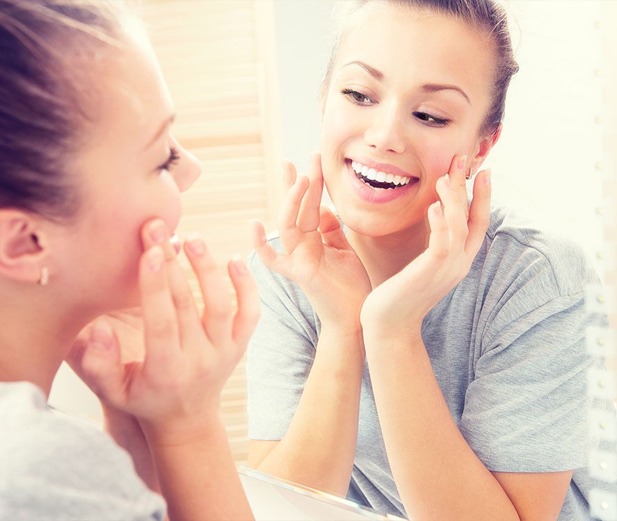 A young woman with a ponytail smiles and touches her face while looking at herself in a mirror.