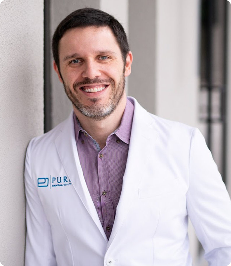 A man with short dark hair and a beard is smiling, wearing a white lab coat over a light purple shirt, standing against a light-colored wall.