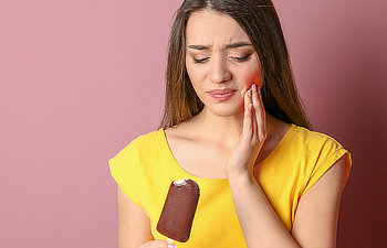 A woman in a yellow shirt holds an ice cream bar and touches her cheek, appearing to feel tooth sensitivity or pain.