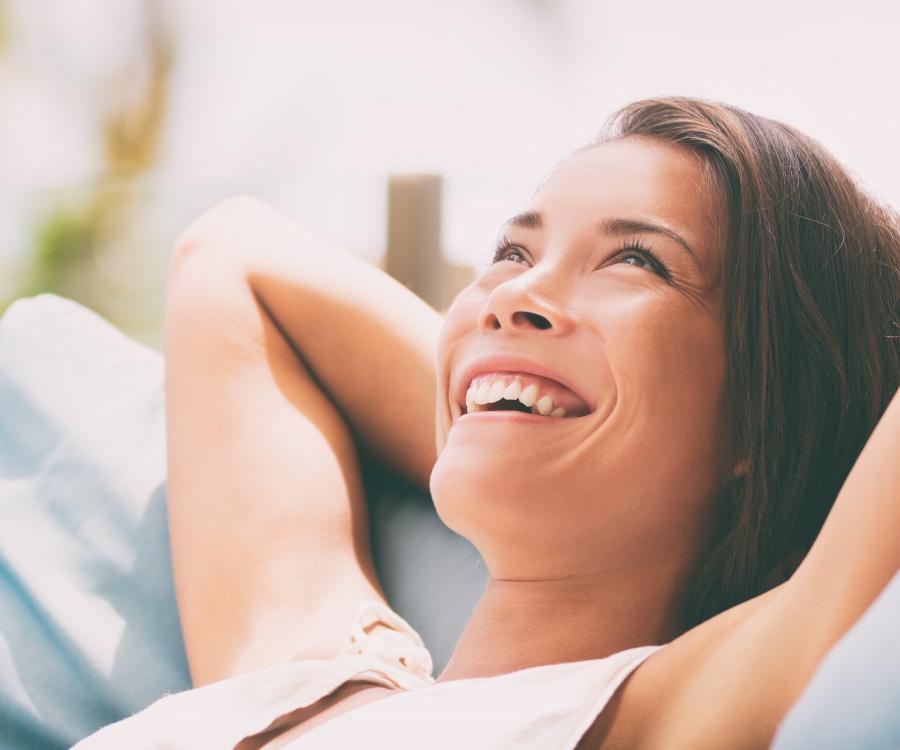A woman reclines outdoors on a blue cushion, smiling and looking upward with her arms resting behind her head.