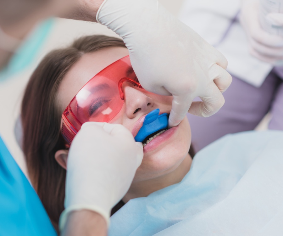 A patient wearing protective glasses and a dental bib receives a blue mouth guard from a gloved dentist during a dental procedure.