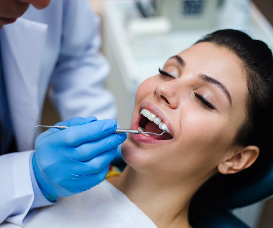 A dentist wearing blue gloves examines a woman's teeth with a dental tool while she sits in a dental chair with her mouth open.