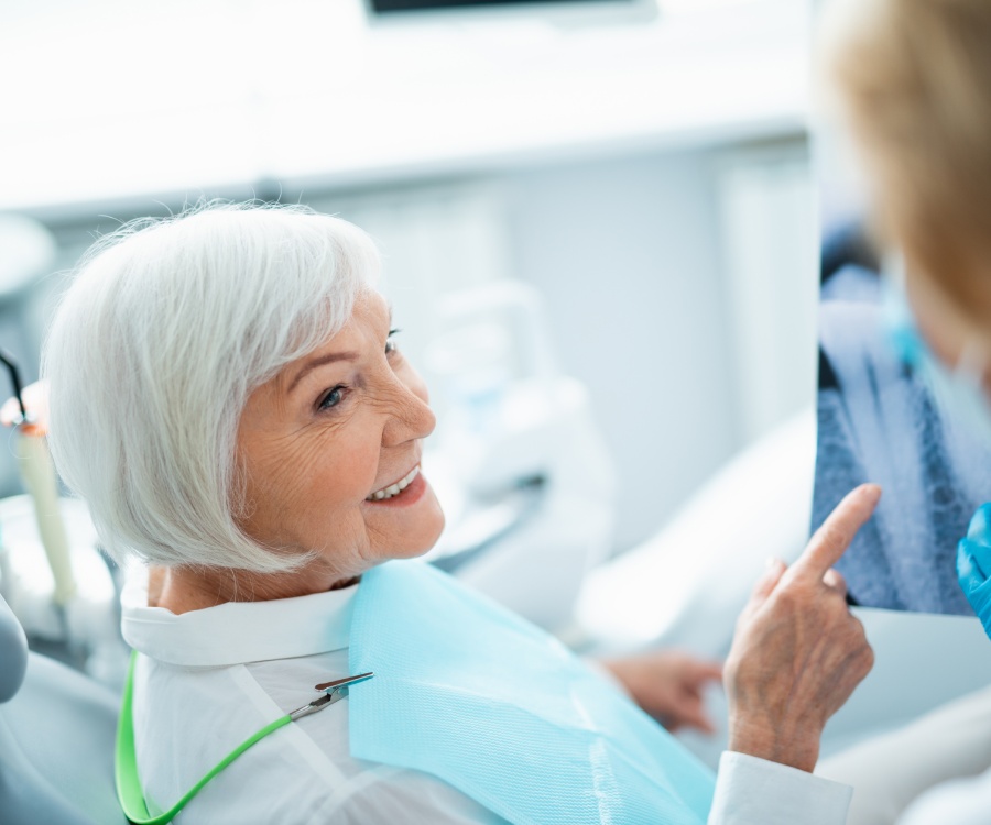 Elderly woman with white hair sitting in a dental chair, smiling and pointing at a dental X-ray image while wearing a blue bib.