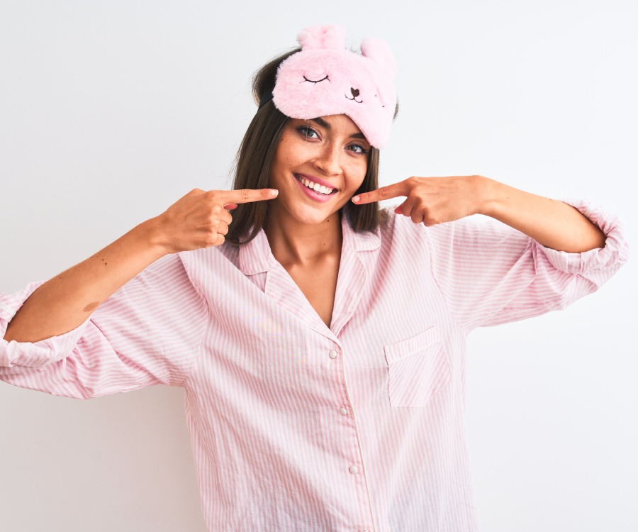 Woman in pink striped pajamas and a pink bunny sleep mask smiles while pointing at her face against a plain white background.