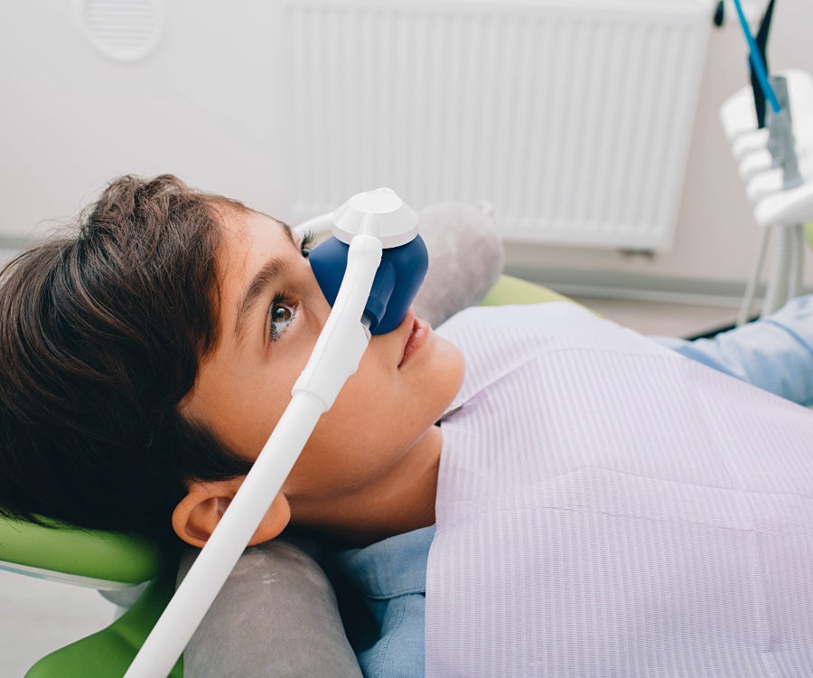 A child is lying on a dental chair wearing a sedation mask over the nose, likely preparing for a dental procedure.