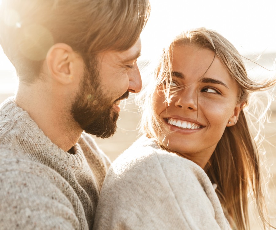 A man and woman wearing sweaters smile at each other outdoors in natural sunlight.