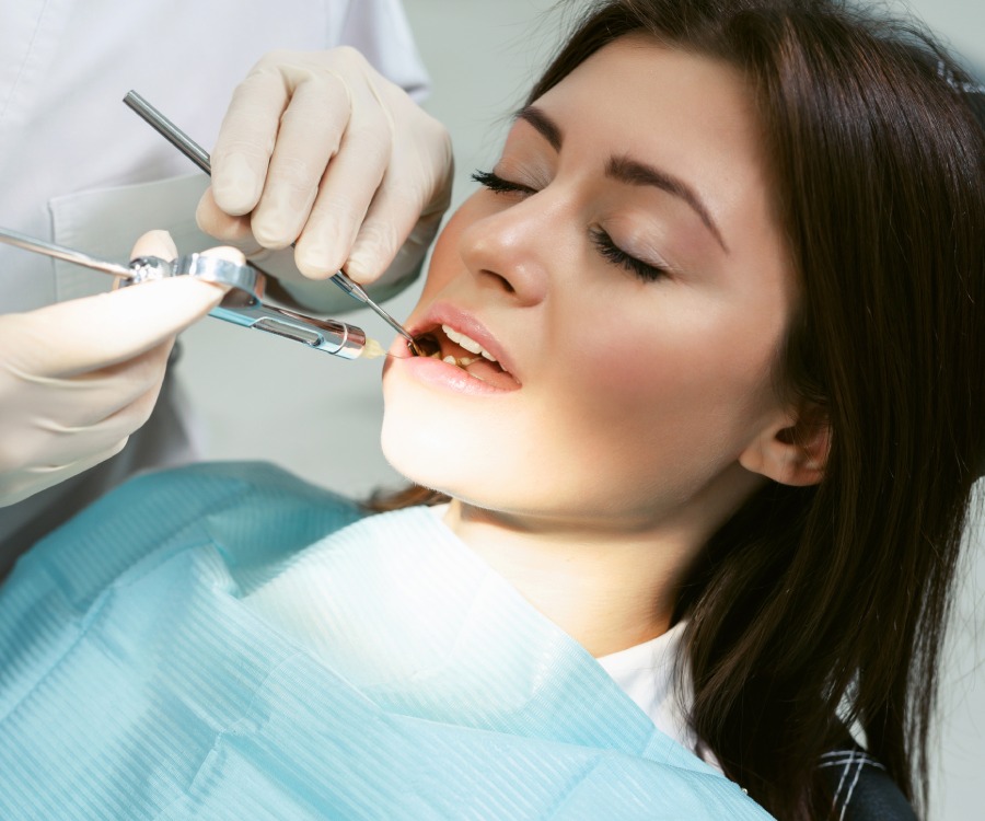A dentist examines a woman's teeth using dental instruments while she sits in the dental chair wearing a blue bib.