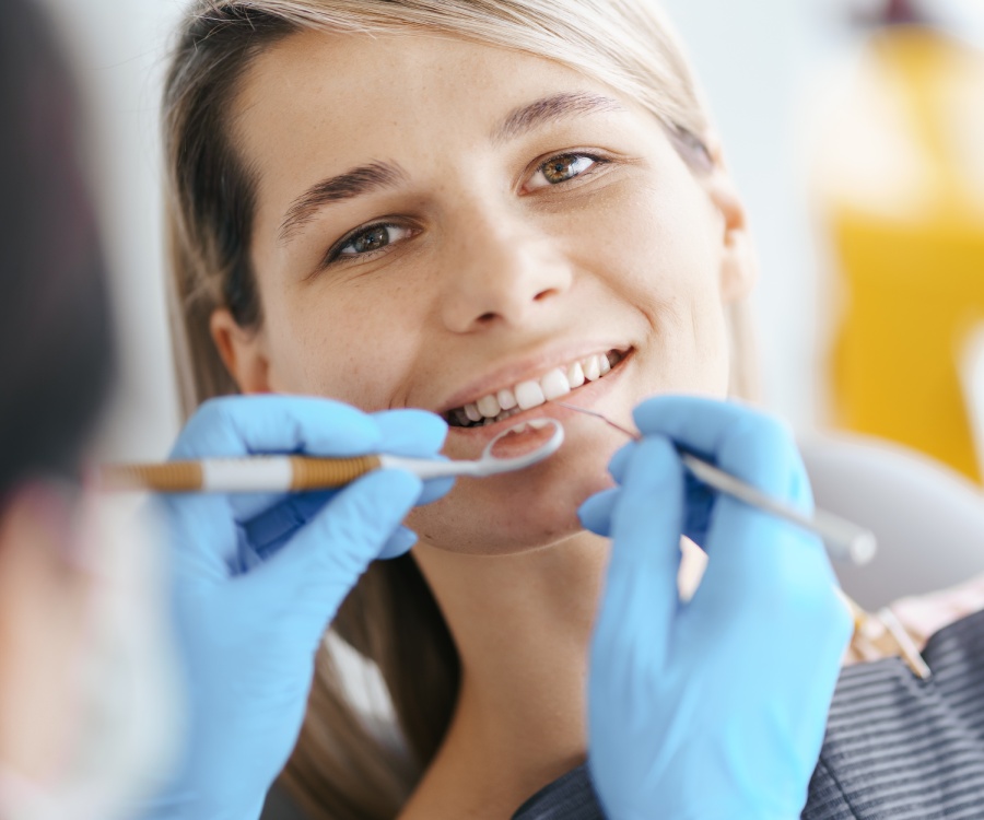 A dentist wearing blue gloves examines a young woman's teeth using dental instruments.