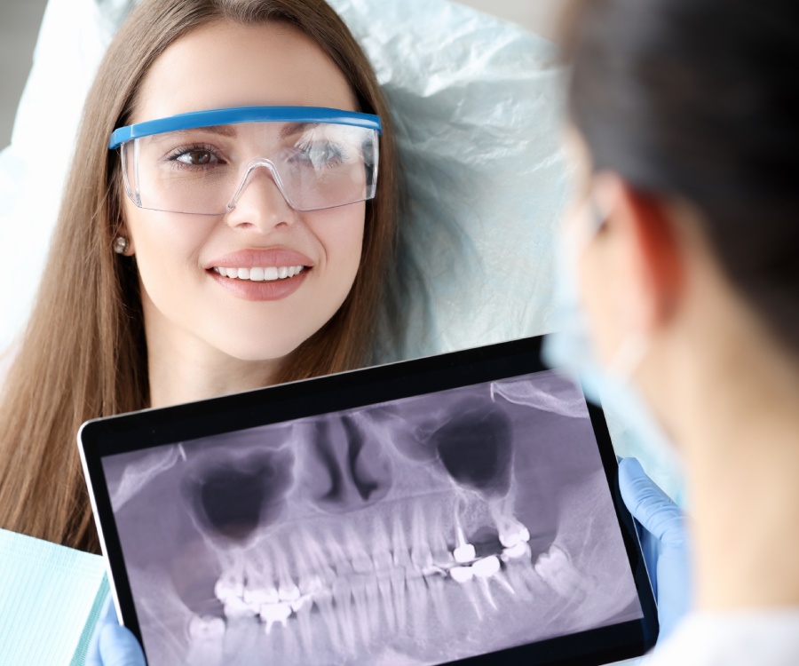 A woman wearing safety glasses sits in a dental chair and smiles at a dentist holding a digital tablet displaying a dental X-ray.
