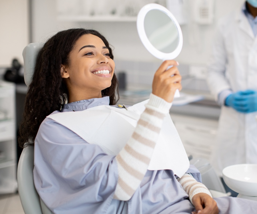 A woman sitting in a dental chair smiles while looking at her reflection in a handheld mirror, with a dentist standing nearby in the background.