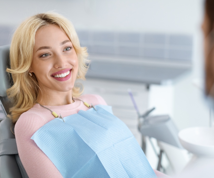 A woman with blonde hair sits in a dental chair, wearing a pink top and a blue dental bib, smiling at someone off-camera.