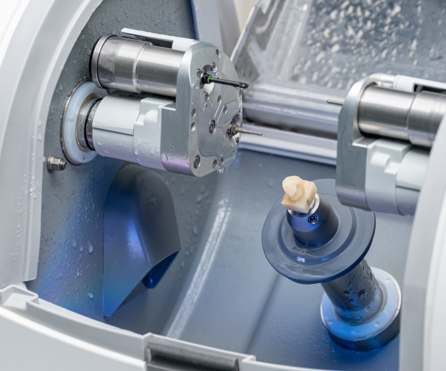 A dental milling machine carving a tooth model from a ceramic block, with the partially milled restoration mounted inside the machine.