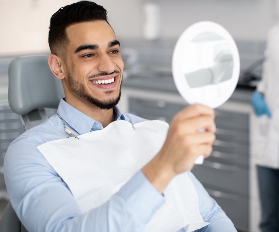 A man sitting in a dental chair smiles while looking at his reflection in a handheld mirror, wearing a dental bib.