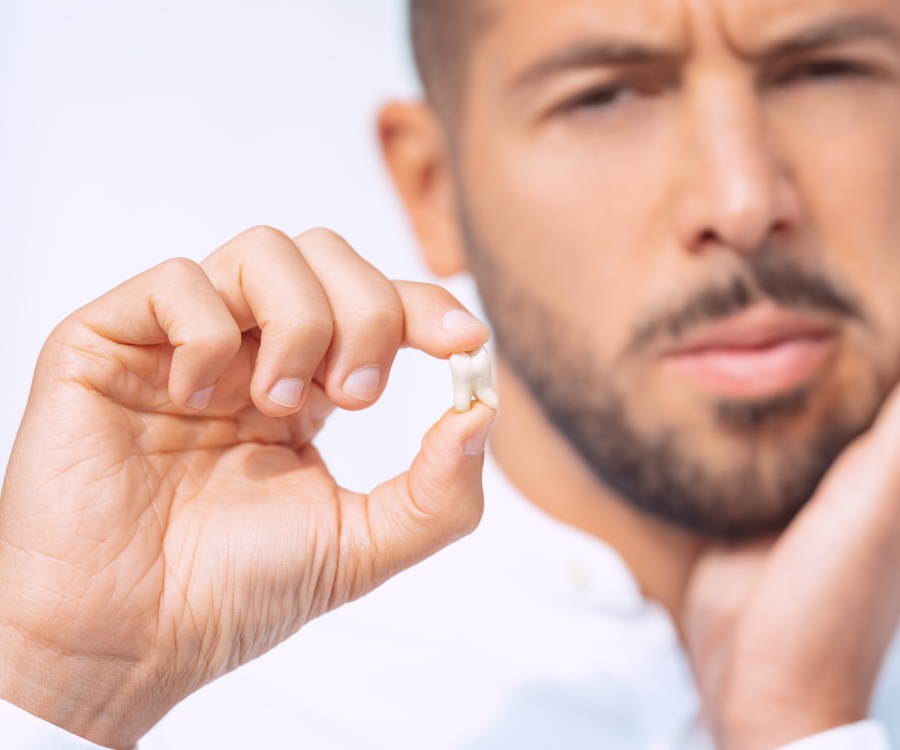 A man holds a tooth in one hand and touches his cheek with the other, suggesting possible dental pain or a recent tooth extraction.