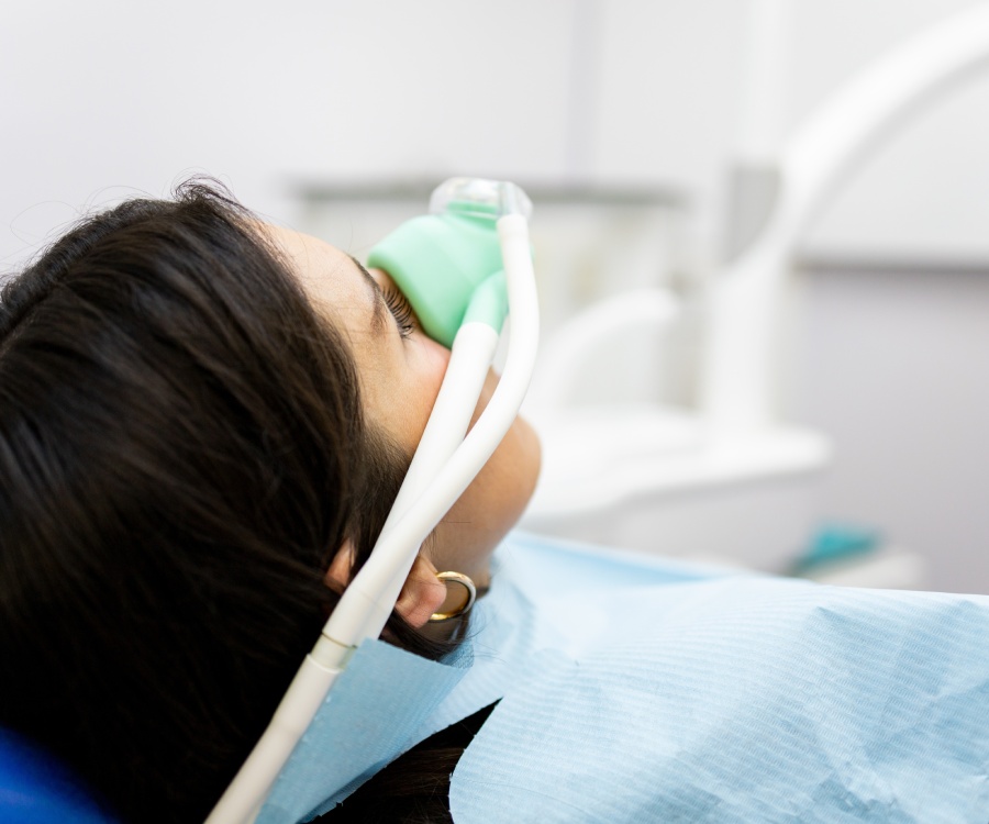 A person lying back in a dental chair wearing a nasal mask for sedation, with dental equipment visible in the background.