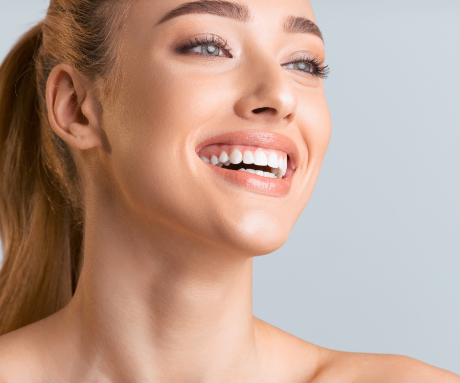 Woman with light skin and brown hair smiling, showing teeth, looking slightly upward to the side against a plain light background.