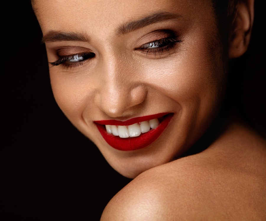 Woman with red lipstick smiling, looking down and to the side, with smooth skin and brown eye makeup against a dark background.