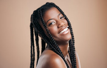 A woman with long braided hair smiles, looking over her shoulder against a plain beige background.