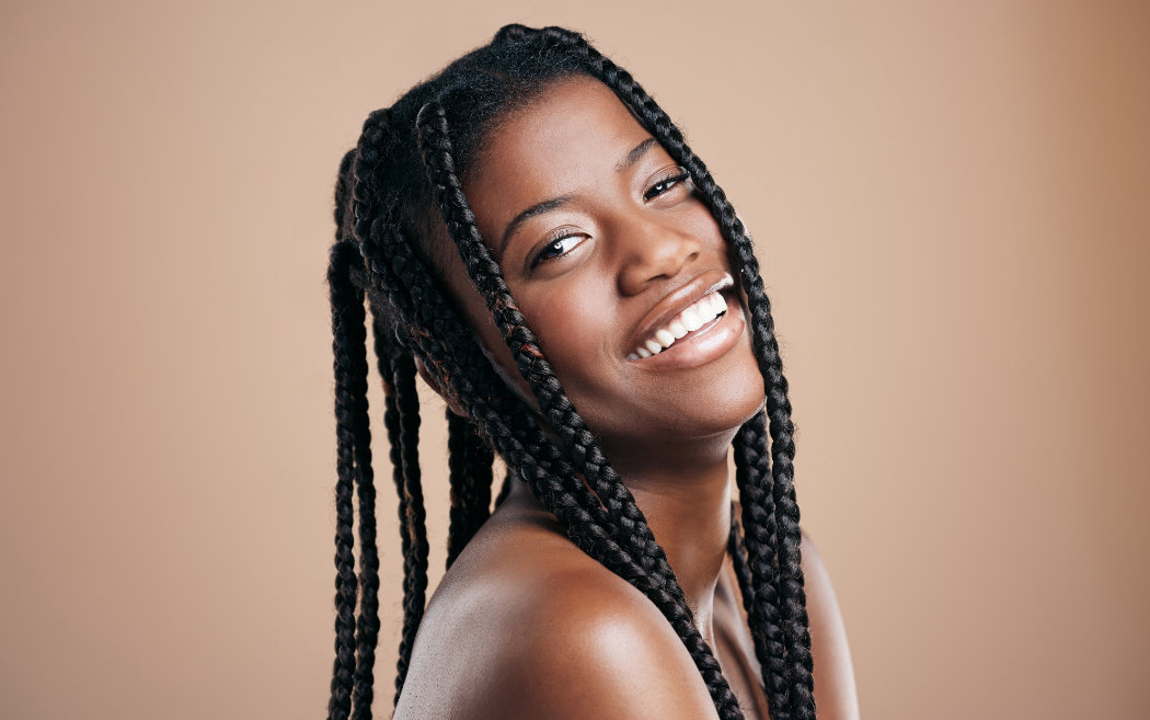 A woman with long braided hair smiles, looking over her shoulder against a plain beige background.