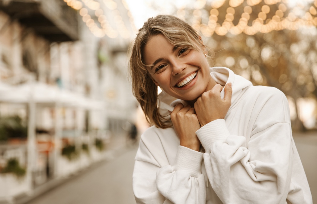 Close up portrait caucasian young happy woman with fresh and clean skin stands outside. Smiling blonde holds collar of white sweatshirt. Lifestyle, female beauty concept