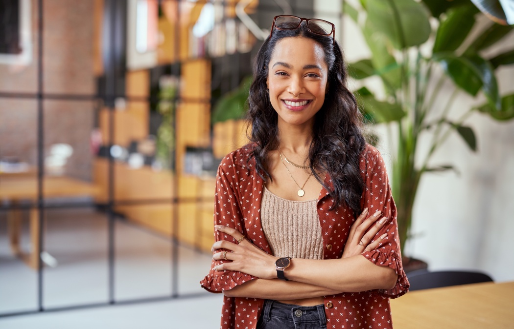 Portrait of smiling young multiethnic woman looking at camera with crossed arms. Successful latin business woman standing in modern office with copy space. Young university hispanic girl with smile.