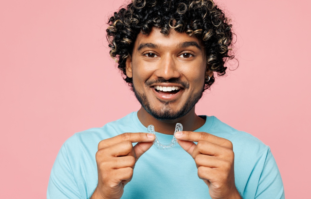Young smiling Indian man he wear blue t-shirt casual clothes hold in hand invisible transparent aligners, invisalign dental braces isolated on plain pastel pink background studio. Lifestyle concept